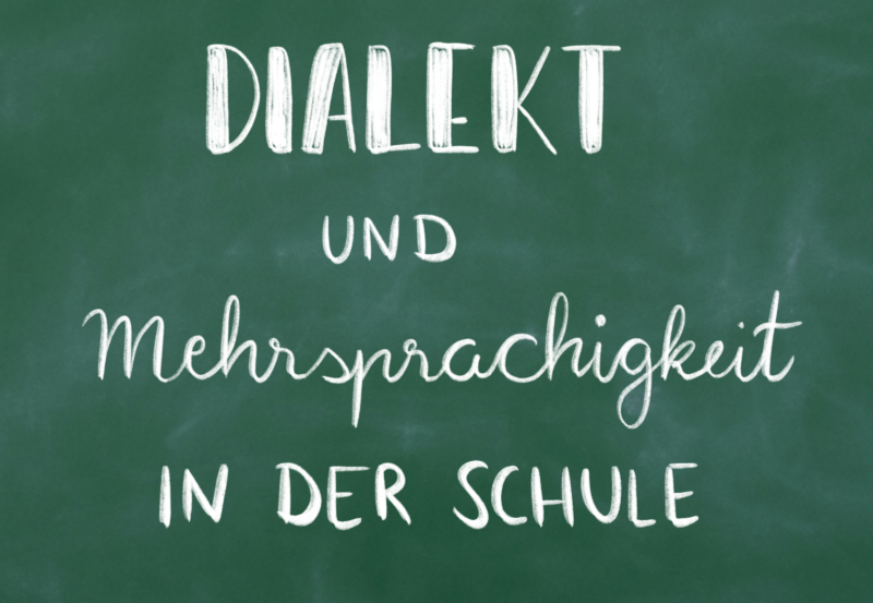 Auf dem Bild ist eine grüne Schultafel zu sehen, auf der mit weißer Kreide "Dialekt und Mehrsprachigkeit in der Schule" geschrieben steht.
